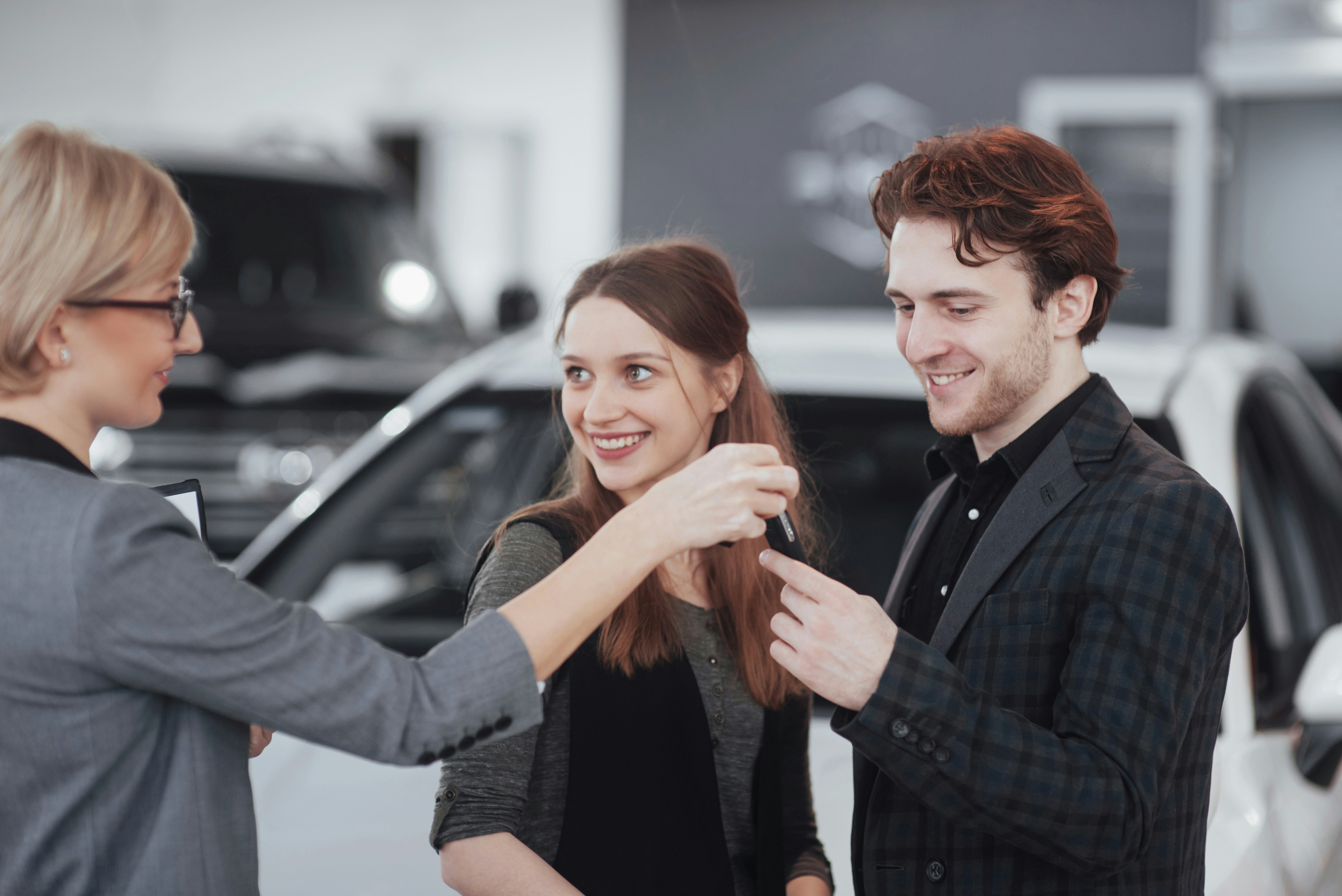Smiling car salesperson handing a set of car keys to a happy young couple inside a dealership, with a white vehicle visible in the background